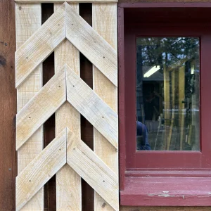 Unfinished pine shutter with open V-shaped chevron slats, hung beside a red-trimmed cabin window