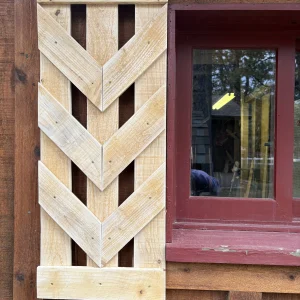Unfinished pine shutter with open V-shaped chevron slats, hung beside a red-trimmed cabin window