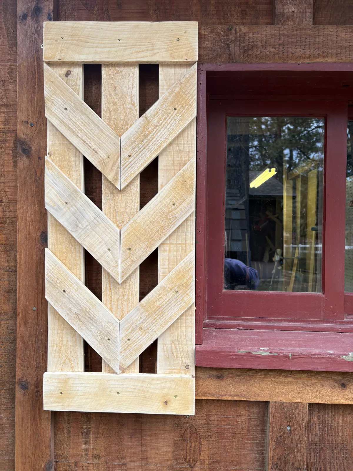 Unfinished pine shutter with open V-shaped chevron slats, hung beside a red-trimmed cabin window
