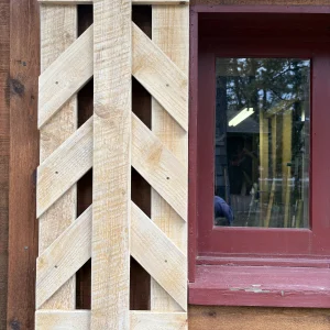 Unfinished pine shutter with a center board and angled chevron slats, hung beside a red-trimmed cabin window