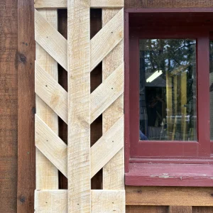 Unfinished pine shutter with a center board and angled chevron slats, hung beside a red-trimmed cabin window