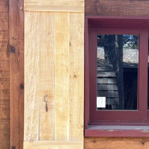 Traditional pine board shutter with top and bottom battens, hung beside a red-trimmed cabin window