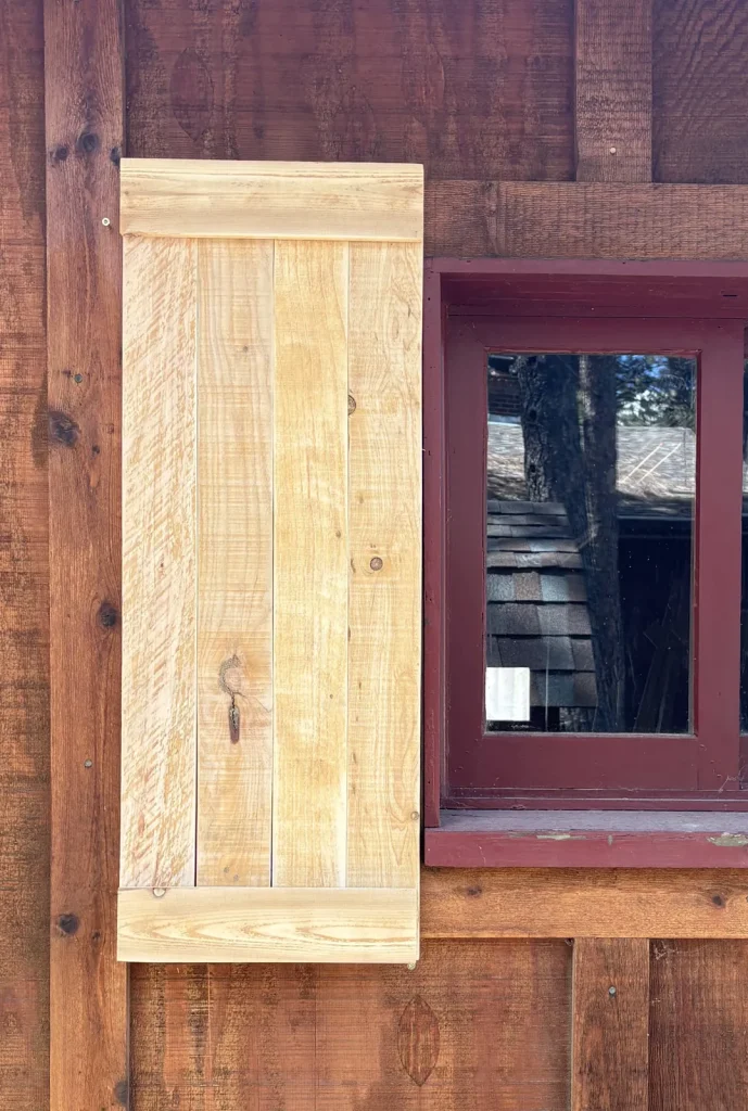Traditional pine board shutter with top and bottom battens, hung beside a red-trimmed cabin window